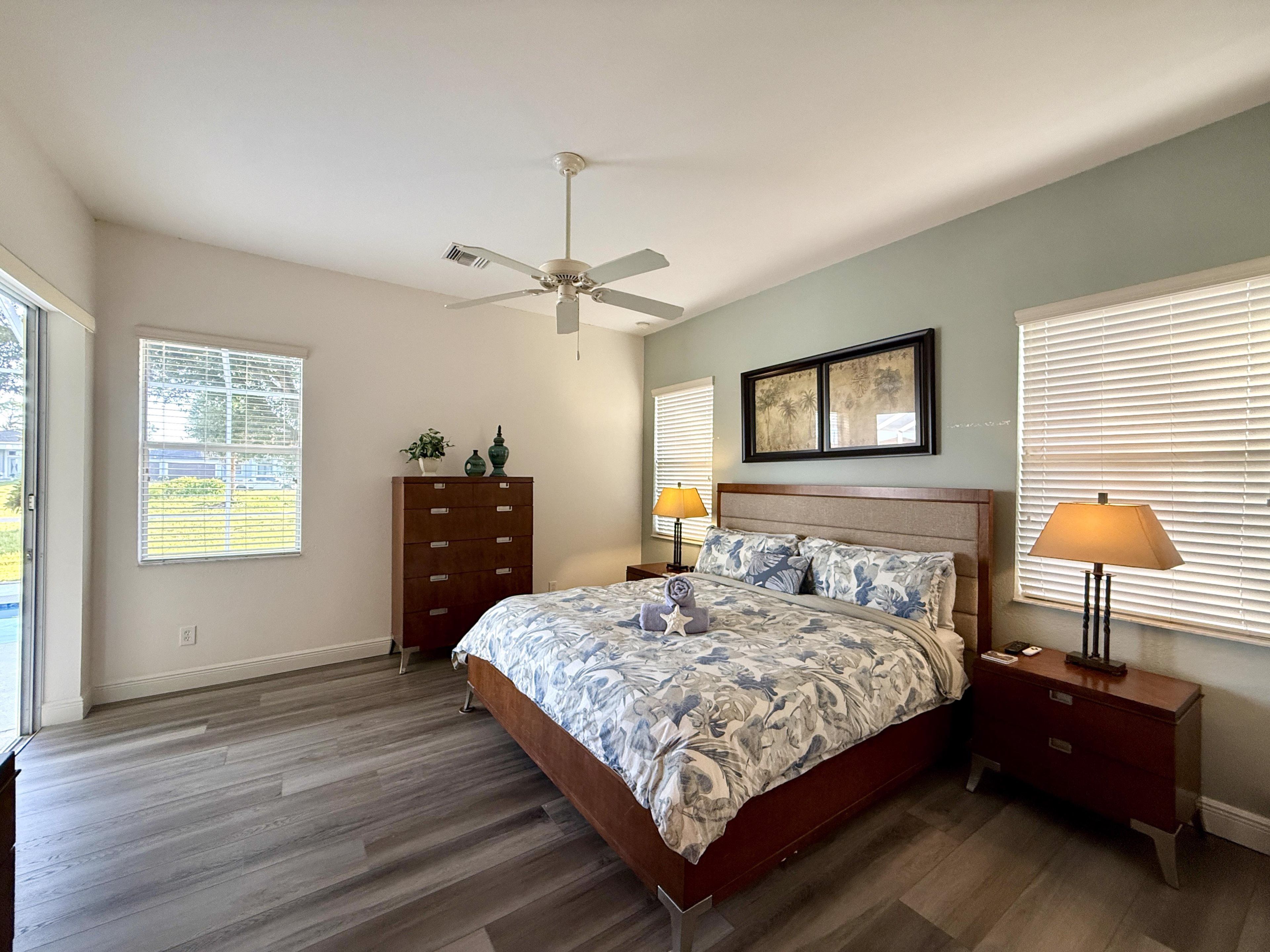 A bedroom with a bed, nightstands, a dresser, a ceiling fan, and large windows with blinds.