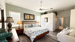 Une chambre à coucher comprenant un lit, deux tables de chevet, un ventilateur de plafond, une fenêtre avec des stores et une porte donnant sur une autre pièce.