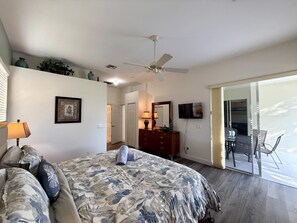 Un dormitorio con una cama, ventilador de techo, televisor y puerta corrediza de vidrio que da a un área exterior.
