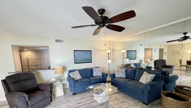 A modern living room with a blue sofa, a dark armchair, a glass coffee table, and a ceiling fan. - Come live the barefoot dream - Your beachside paradise, and the ocean is your backyard! (Englewood)
