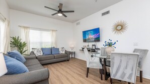 A modern living room with a gray sofa, a dining table with chairs, a ceiling fan, and a TV on the wall.