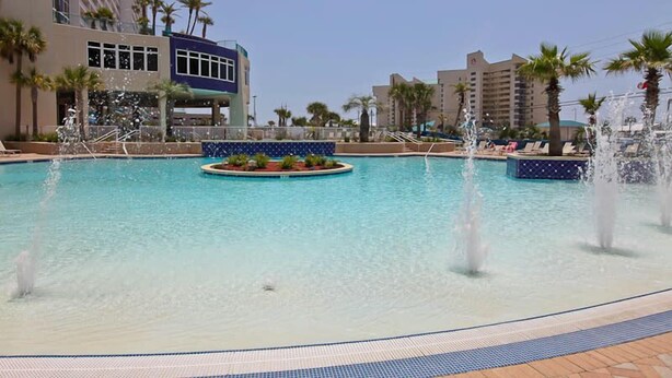 Una piscina grande al aire libre con múltiples chorros de agua, rodeada de palmeras y edificios modernos.