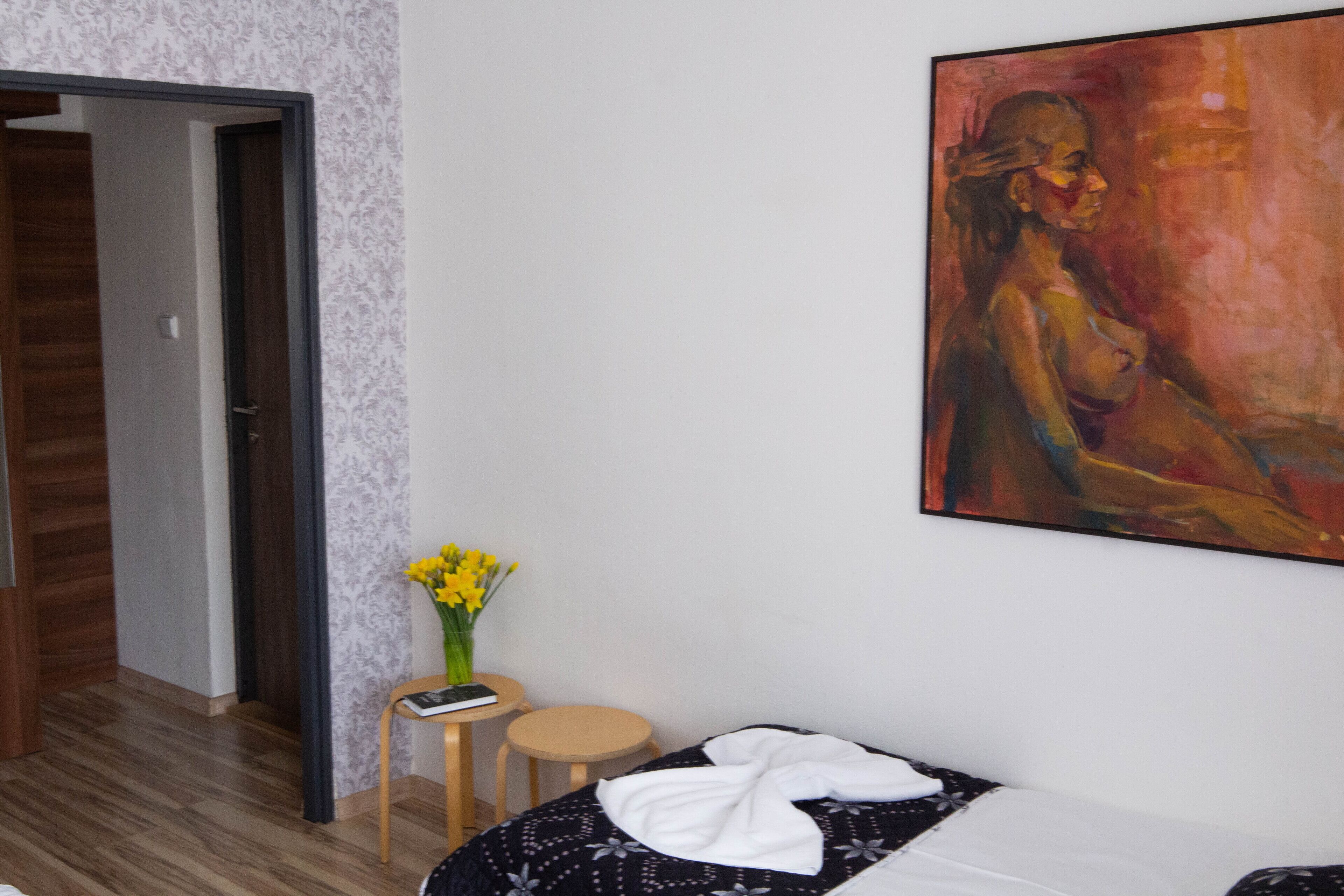Una habitación de hotel con una cama, una mesita con un jarrón de flores, un cuadro en la pared y una puerta que da a otra habitación.