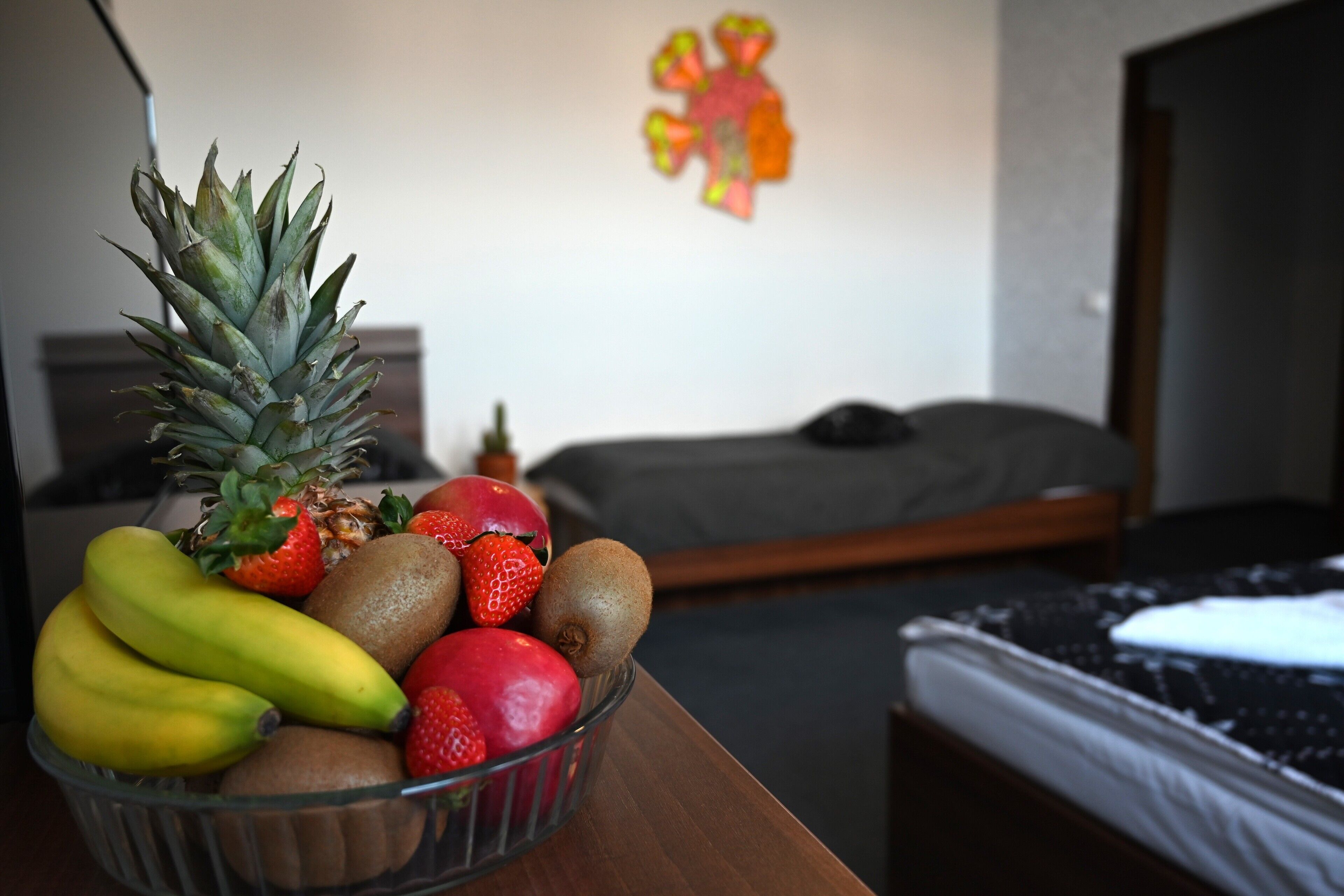 A room with a bed, a bedside table with a fruit bowl, and a wall decoration.