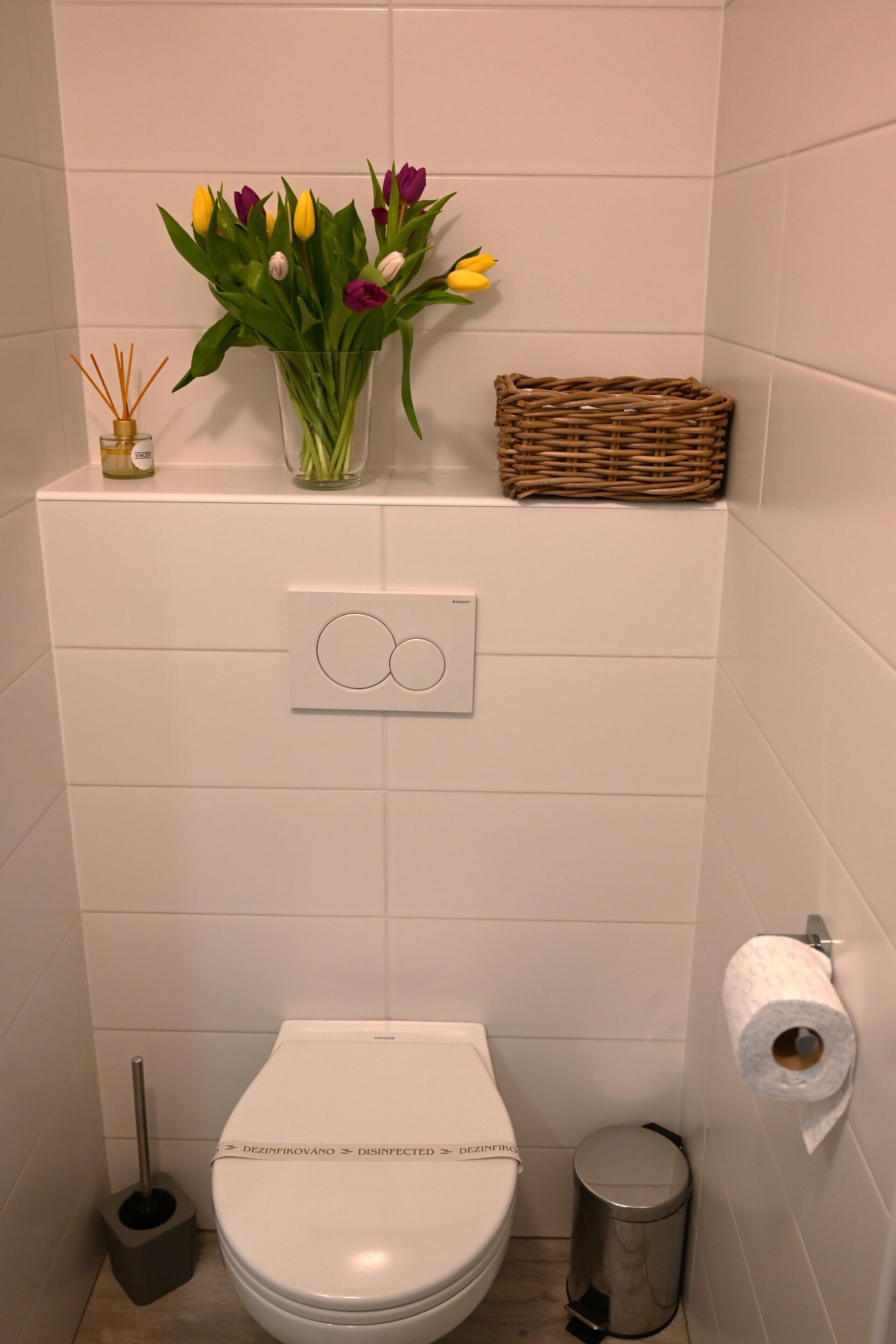 Une salle de bain comprenant des toilettes, une poubelle, un porte-rouleau de papier toilette, un diffuseur de parfum à bâtonnets et un panier en osier posé sur une étagère.