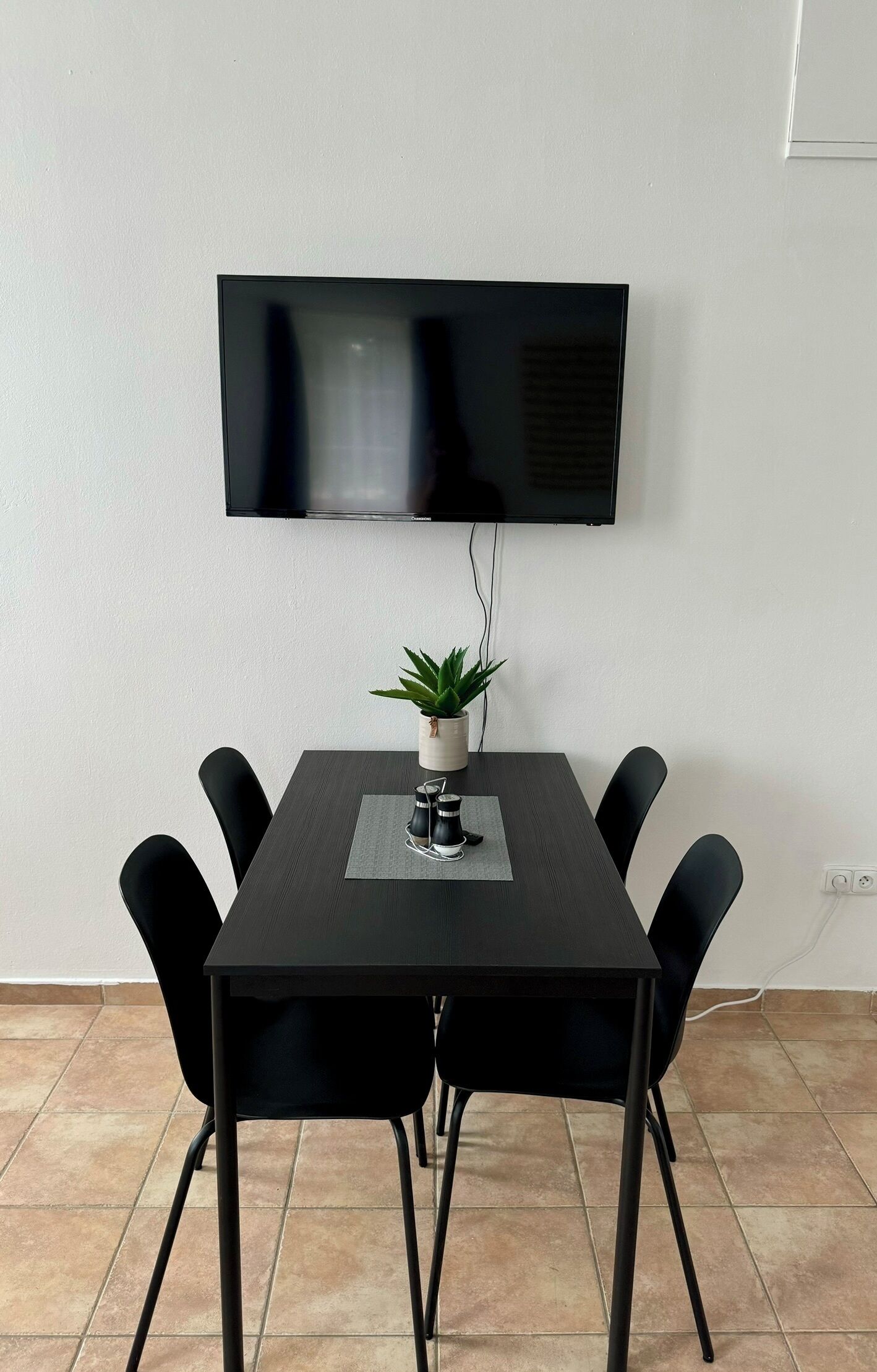 A dining area with a dark wooden table, six black chairs, a potted plant, and a flat-screen TV mounted on the wall.