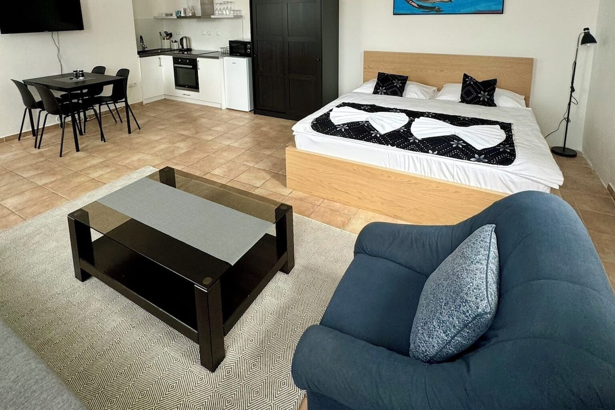A modern hotel room with a bed, a sofa, a dining table, and a kitchenette.