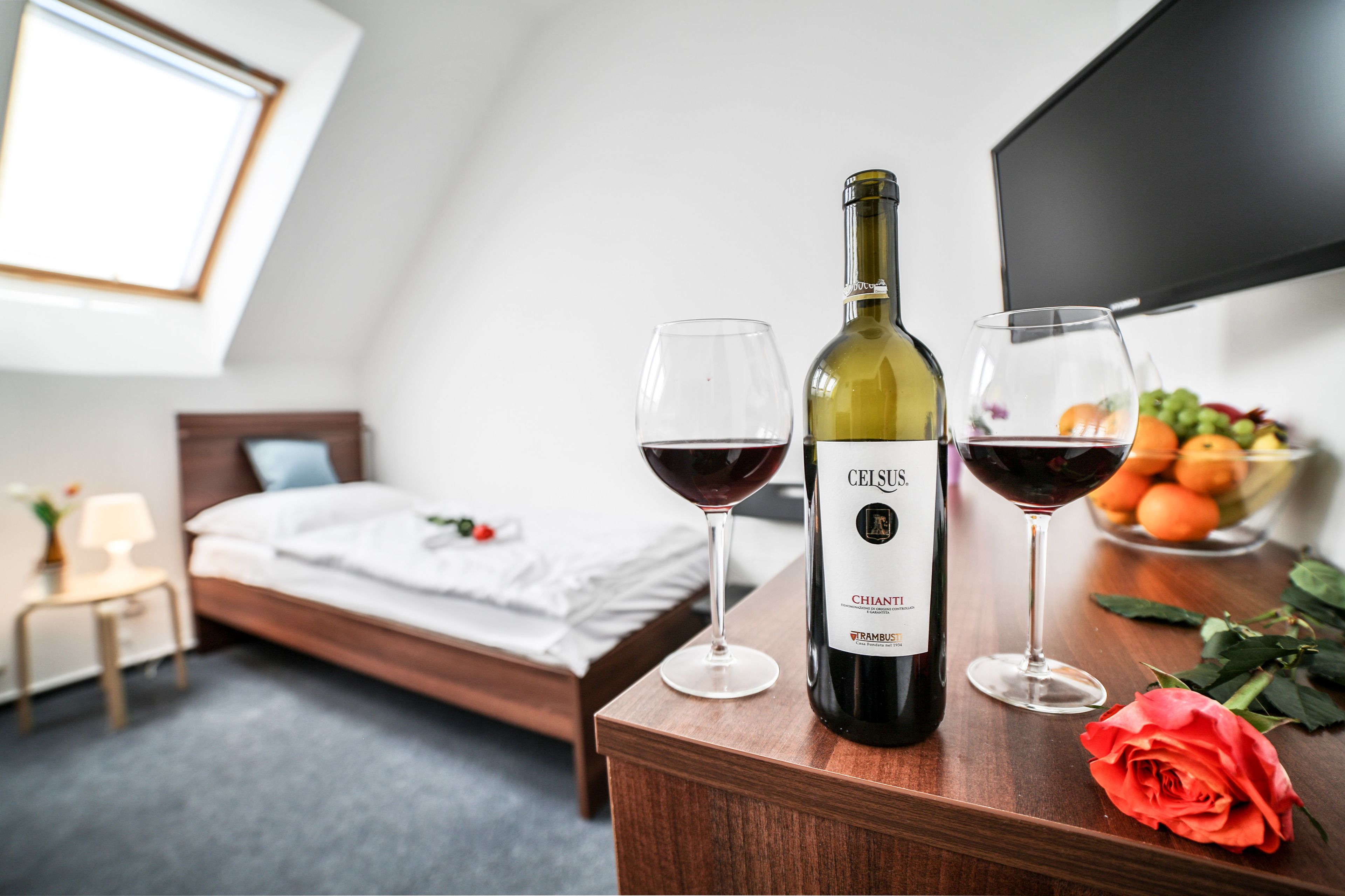 A hotel room with a bed, a wooden nightstand, a bottle of wine, two wine glasses, a rose, and a bowl of fruit.