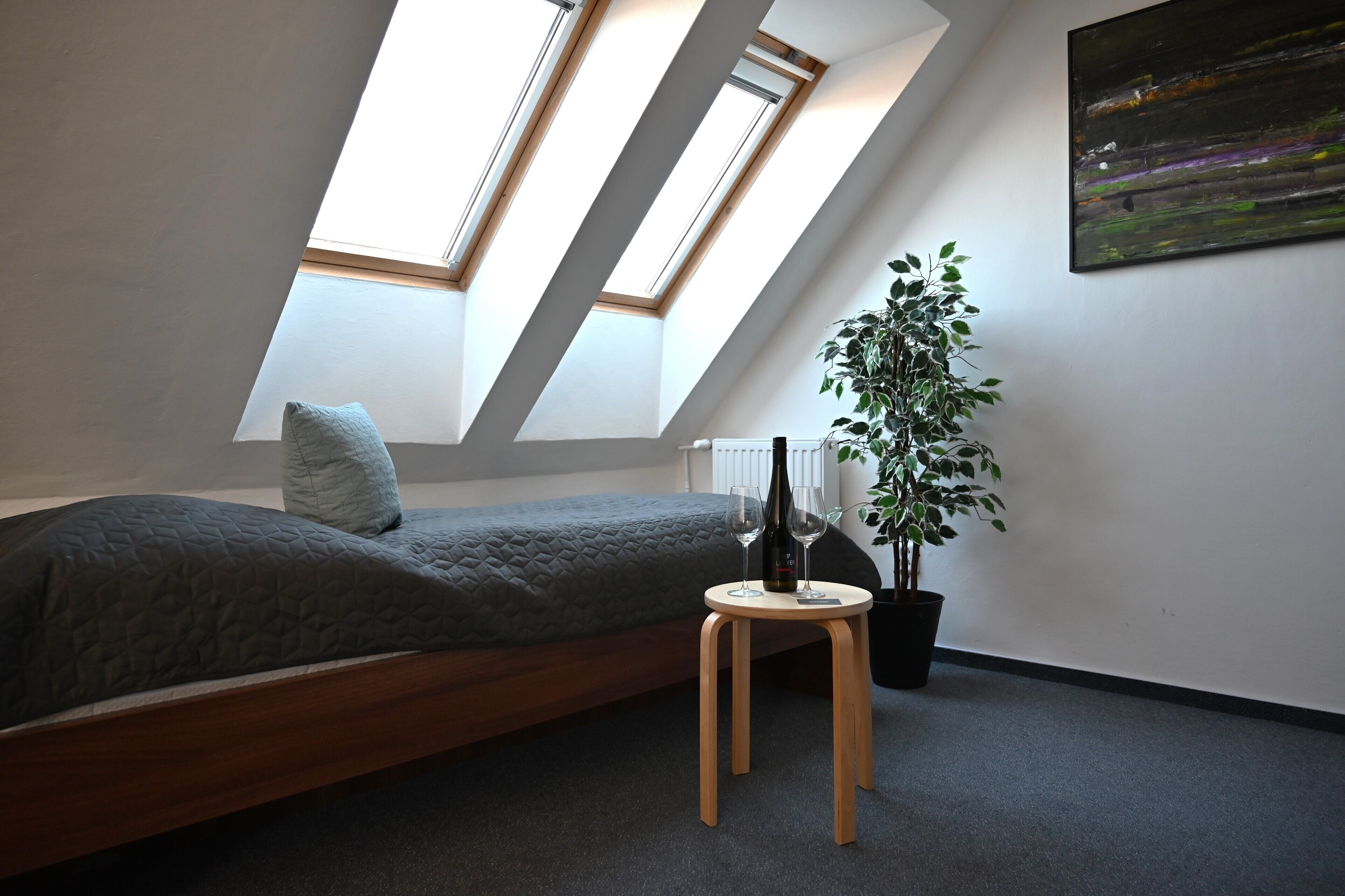Una camera da letto con un soffitto spiovente, lucernari, un letto, uno sgabello di legno, una pianta in vaso e un quadro appeso al muro.