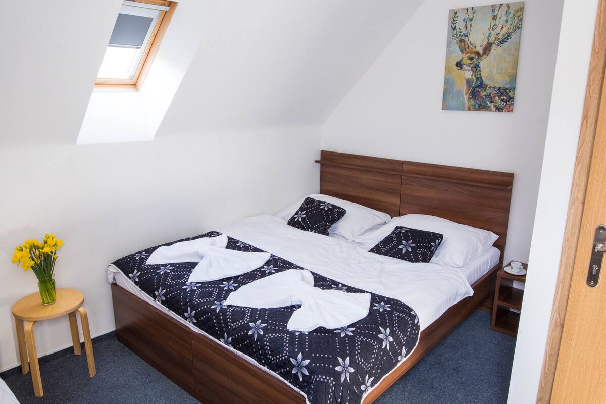 A neatly made bed with a patterned bedspread, a wooden headboard, a small bedside table with a cup, and a vase with flowers on a stool.