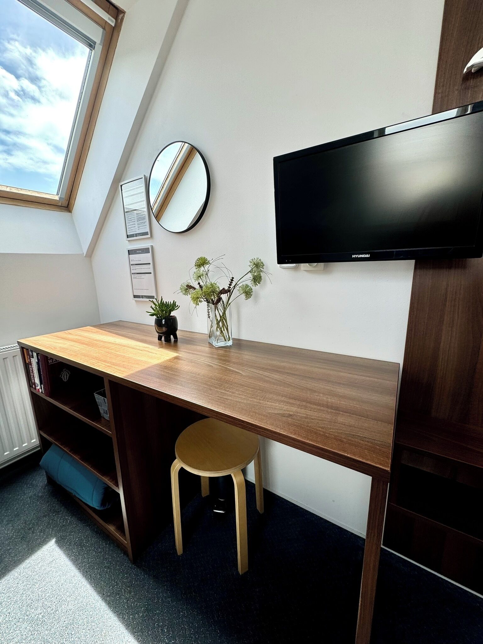 A modern study room with a slanted ceiling, a wooden desk, a chair, a TV, a mirror, and a small potted plant.
