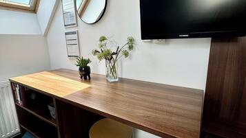 A modern study room with a slanted ceiling, a wooden desk, a chair, a TV, a mirror, and a small potted plant.