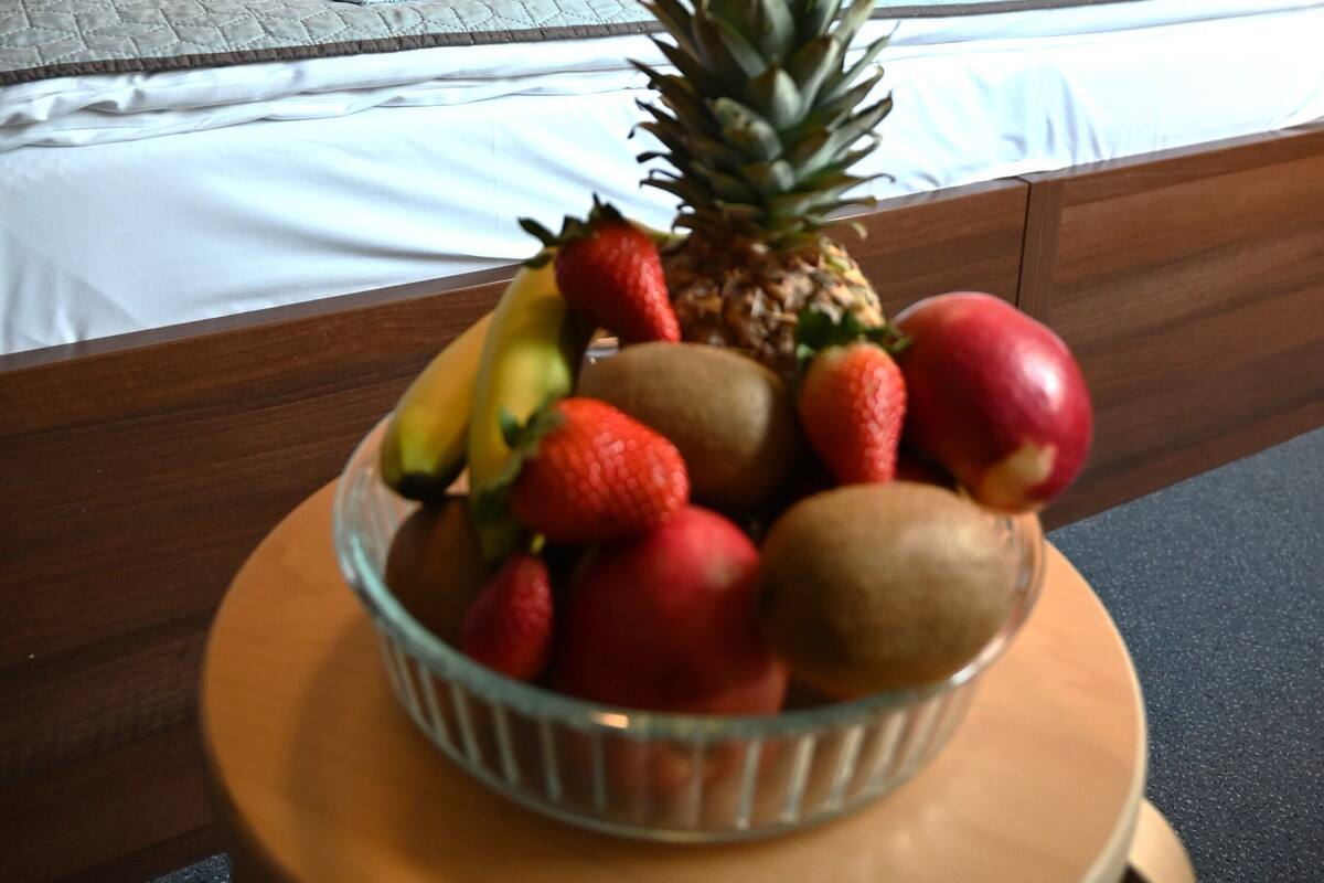 A hotel room with a neatly made bed, a bowl of fruit on a side table, and a patterned wallpaper on the wall.