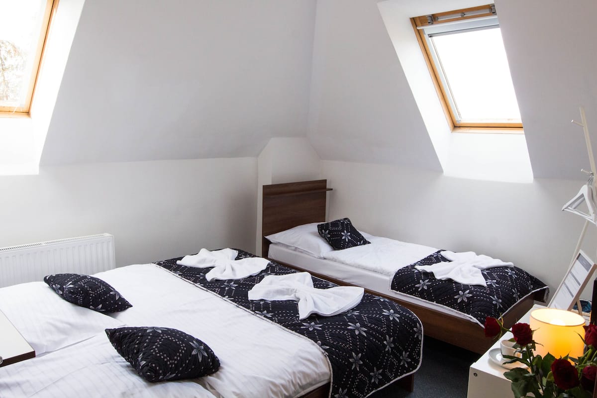 A room with two beds, a radiator, a window, and a bedside table with a lamp and flowers.