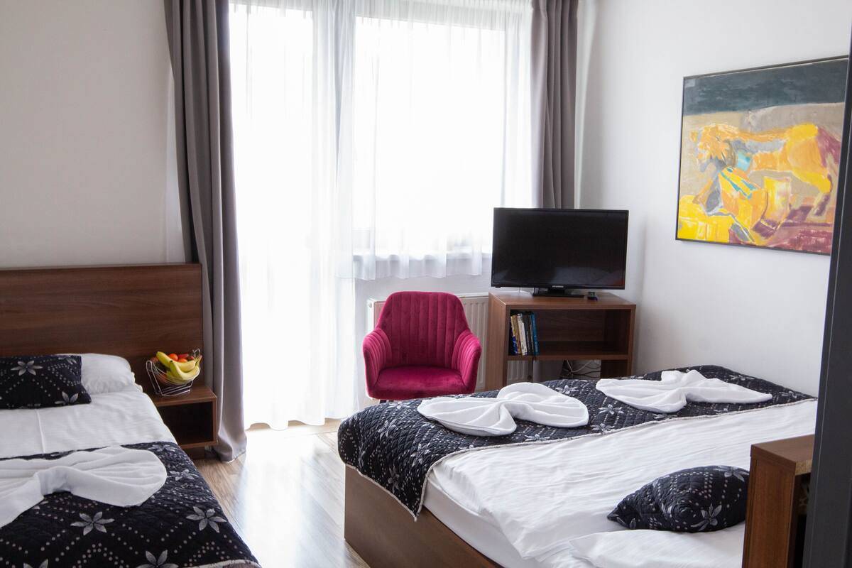 A hotel room with two beds, a TV, a red armchair, and a painting on the wall.