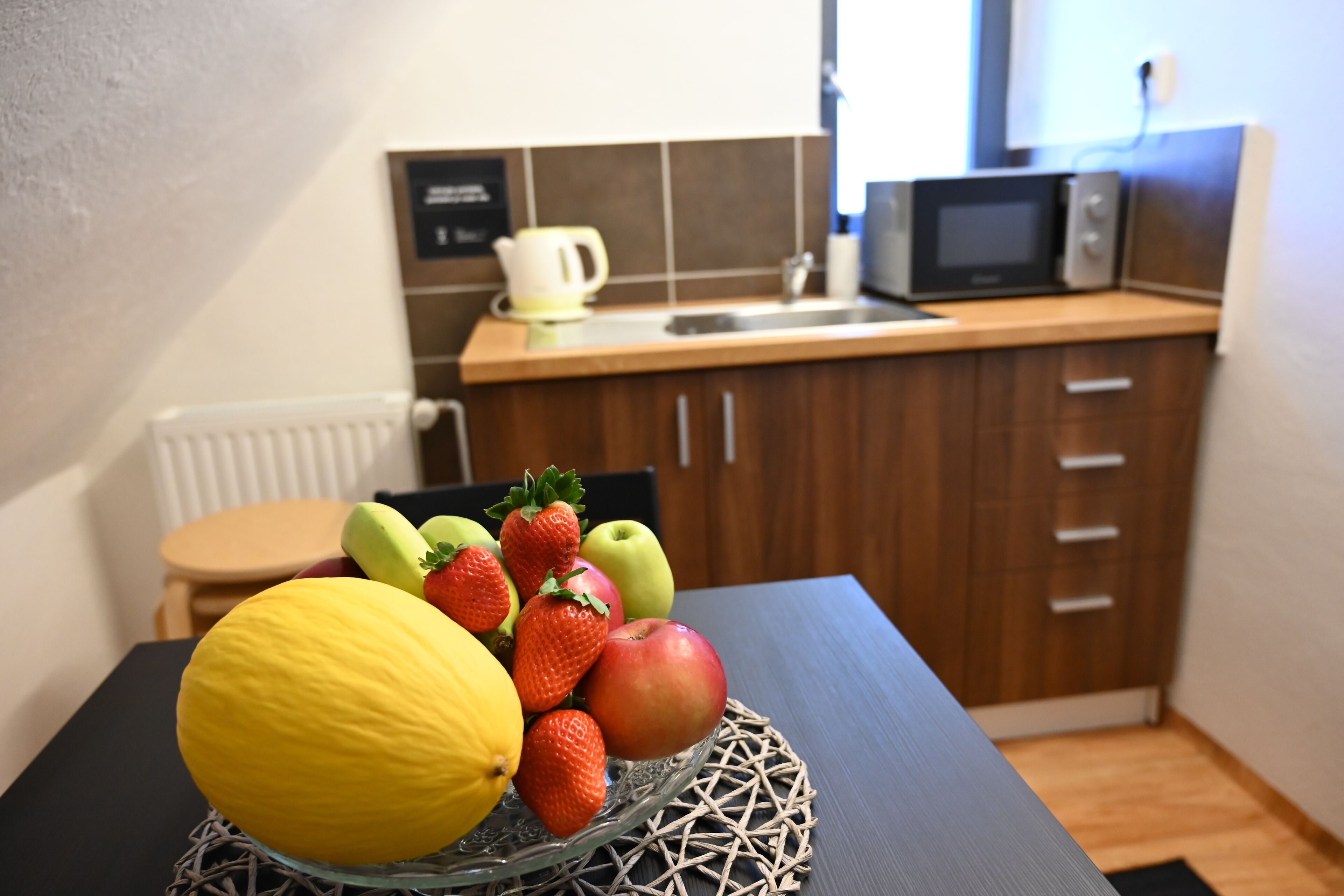 A small kitchen with a microwave, kettle, and wooden cabinets. A fruit bowl with a lemon, apple, and strawberries is on the table.