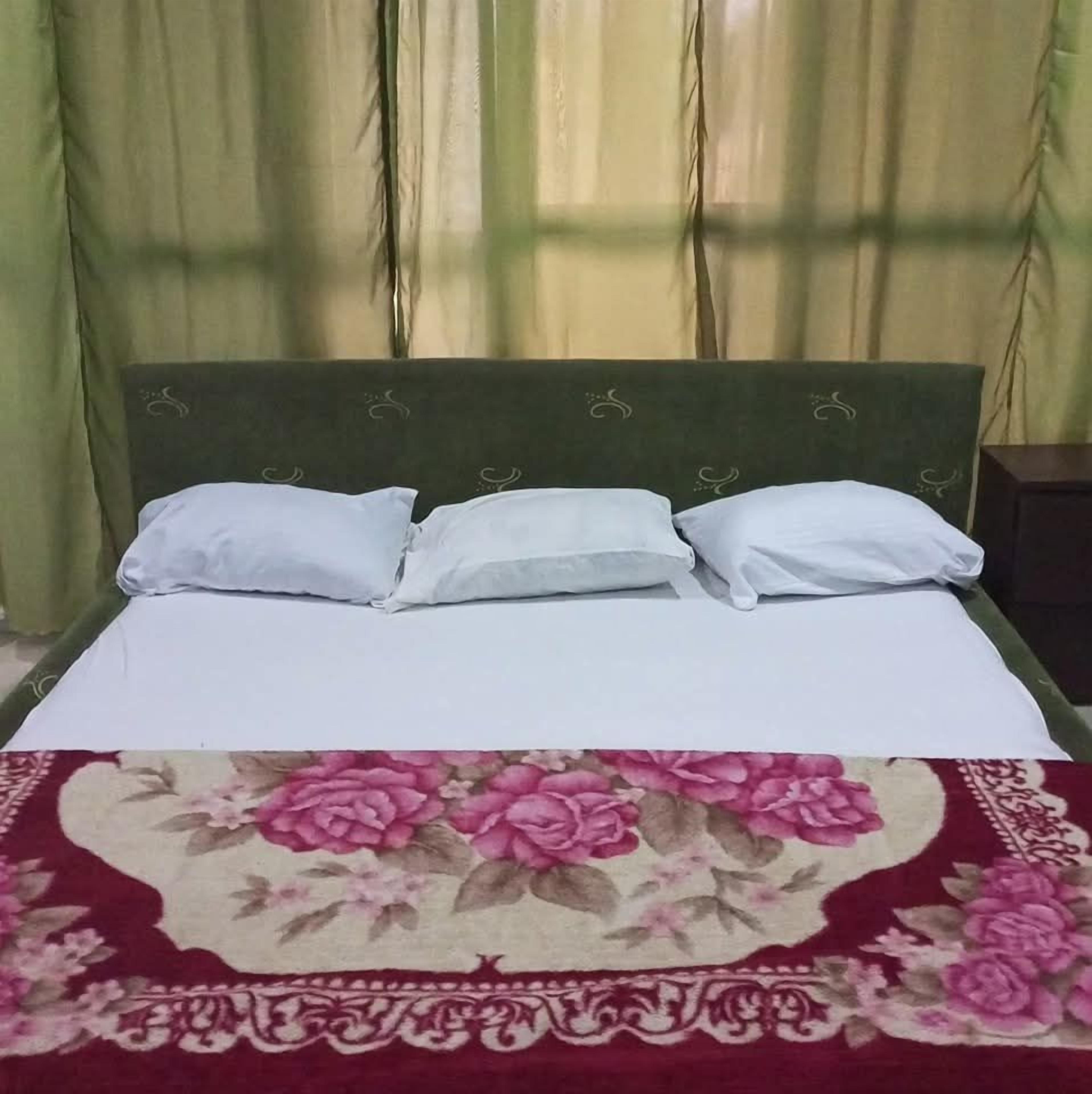 Una cama con cabecera tapizada en verde, ropa de cama blanca y una alfombra con estampado floral.