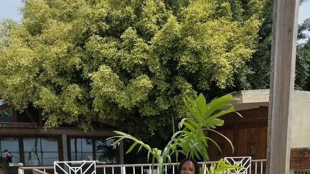 A person sitting on a concrete surface near a white planter with a large plant.