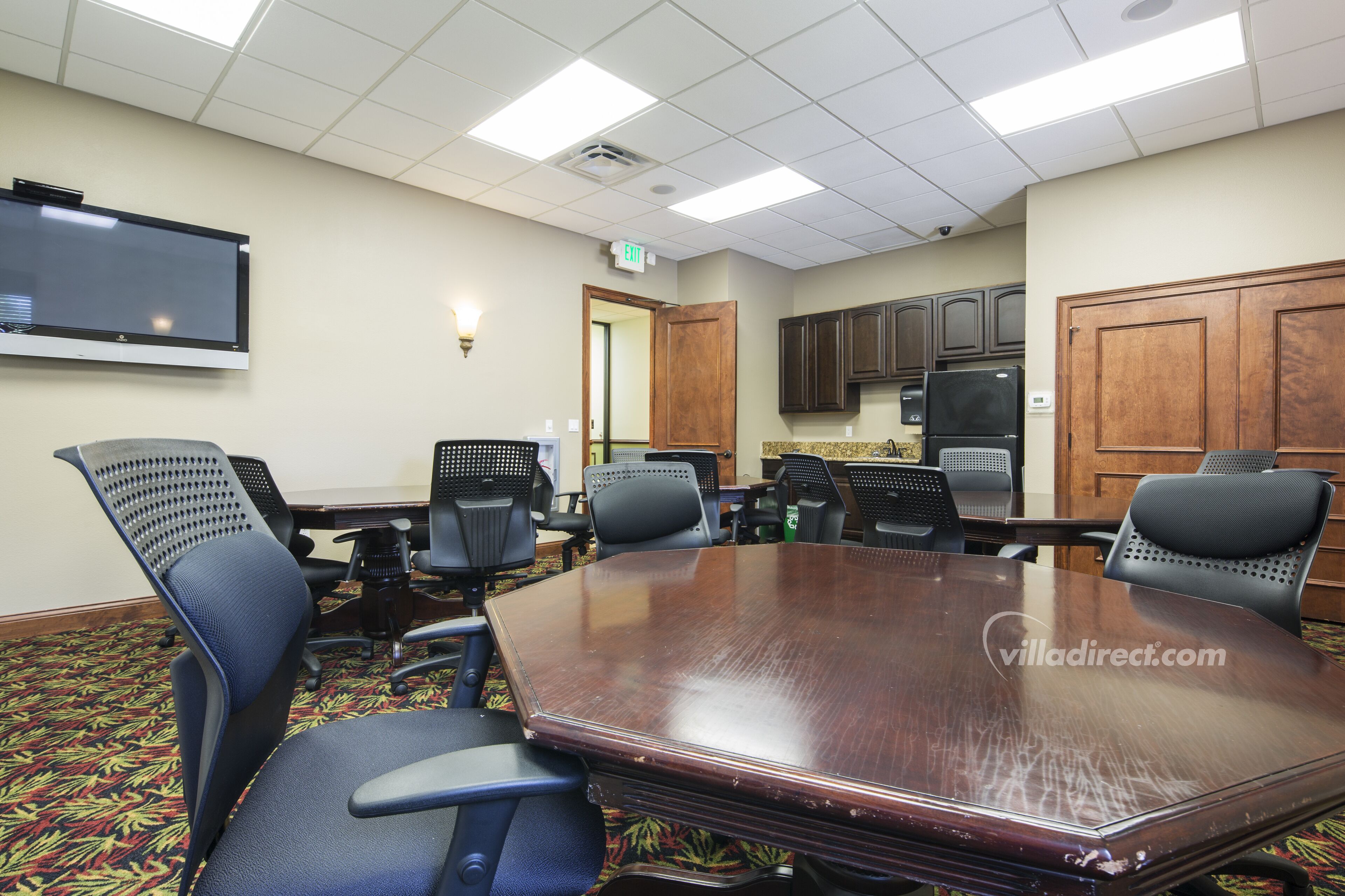 A conference room with a large wooden table, six chairs, a flat-screen TV, and a kitchenette area.