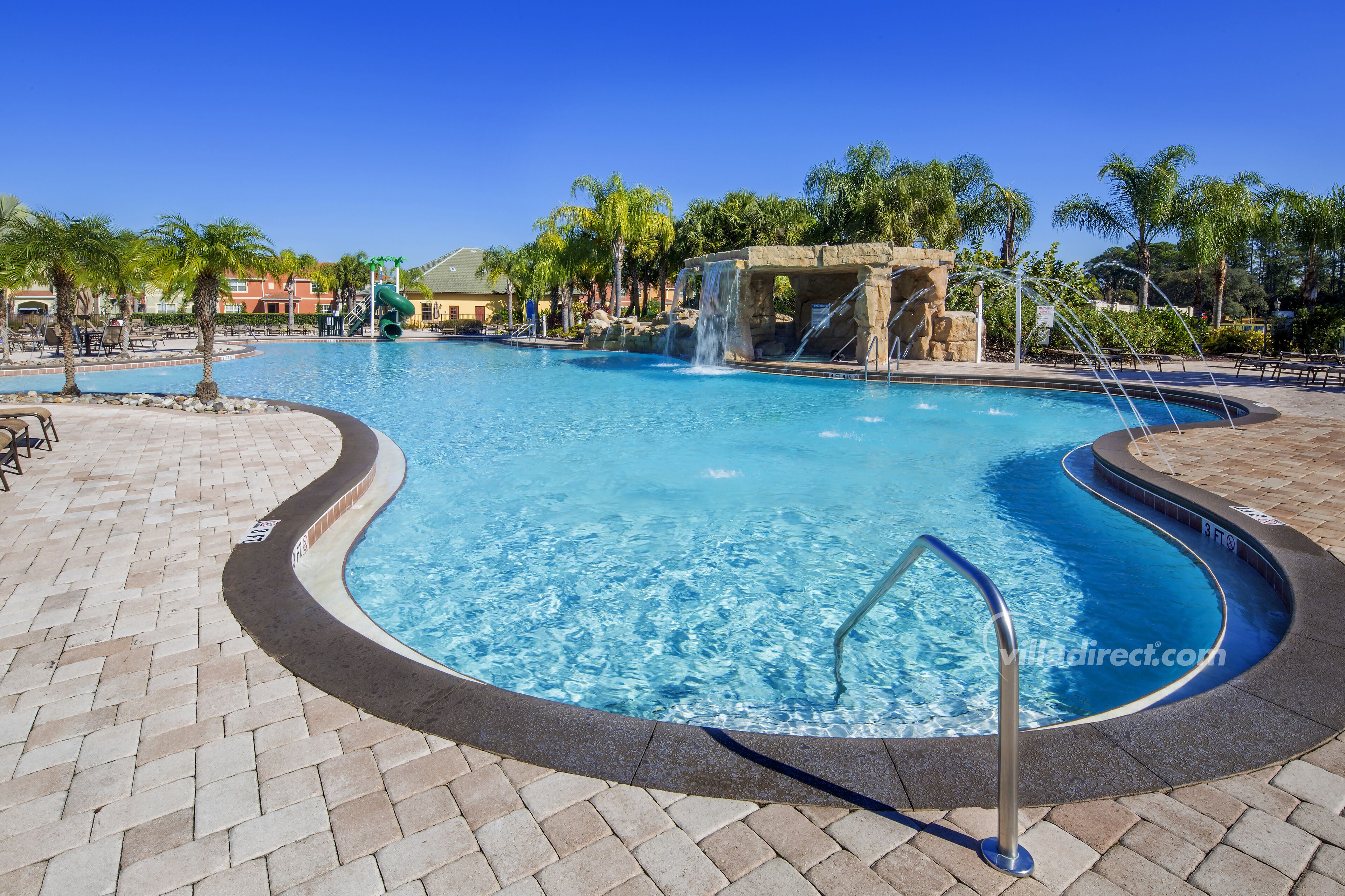 A swimming pool with a waterfall feature, surrounded by palm trees and a brick walkway.