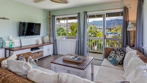 A living room with a TV, a ceiling fan, a sofa, a coffee table, and a view of a balcony with a scenic landscape. - Relax at Kapakai Cottage – 2-Bedroom Comfort with Full Kitchen & W/D (Kailua)