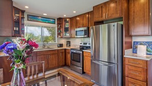 A modern kitchen with wooden cabinets, stainless steel appliances, and a dining area with a glass table. - Relax at Kapakai Cottage – 2-Bedroom Comfort with Full Kitchen & W/D (Kailua)