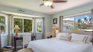 A bedroom with a bed, two bedside tables, a desk, a chair, a ceiling fan, and large windows with a view of palm trees and a house. - Relax at Kapakai Cottage – 2-Bedroom Comfort with Full Kitchen & W/D (Kailua)