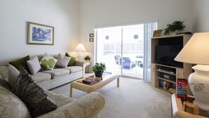 Un salon avec un canapé, une table basse et un téléviseur. On y trouve une plante posée sur une étagère et une fenêtre donnant sur l’extérieur.