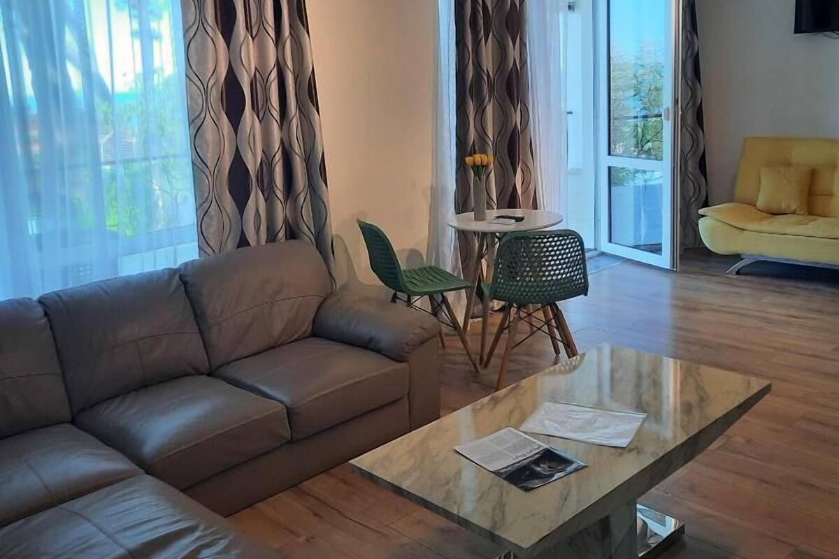A modern living room with a sofa, a glass coffee table, a dining area with chairs, and a yellow armchair.