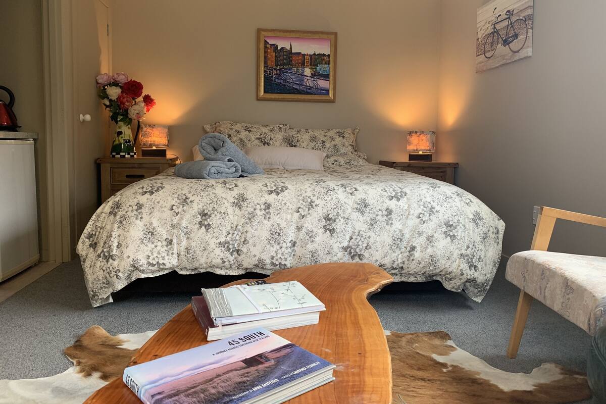 A bedroom with a floral bedspread, a framed cityscape painting, a wooden bedside table with books, a cowhide rug, and a small table with a book.