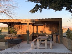 Una zona de barbacoa rústica al aire libre con una estructura de madera, una parrilla de metal, una mesa y un banco.