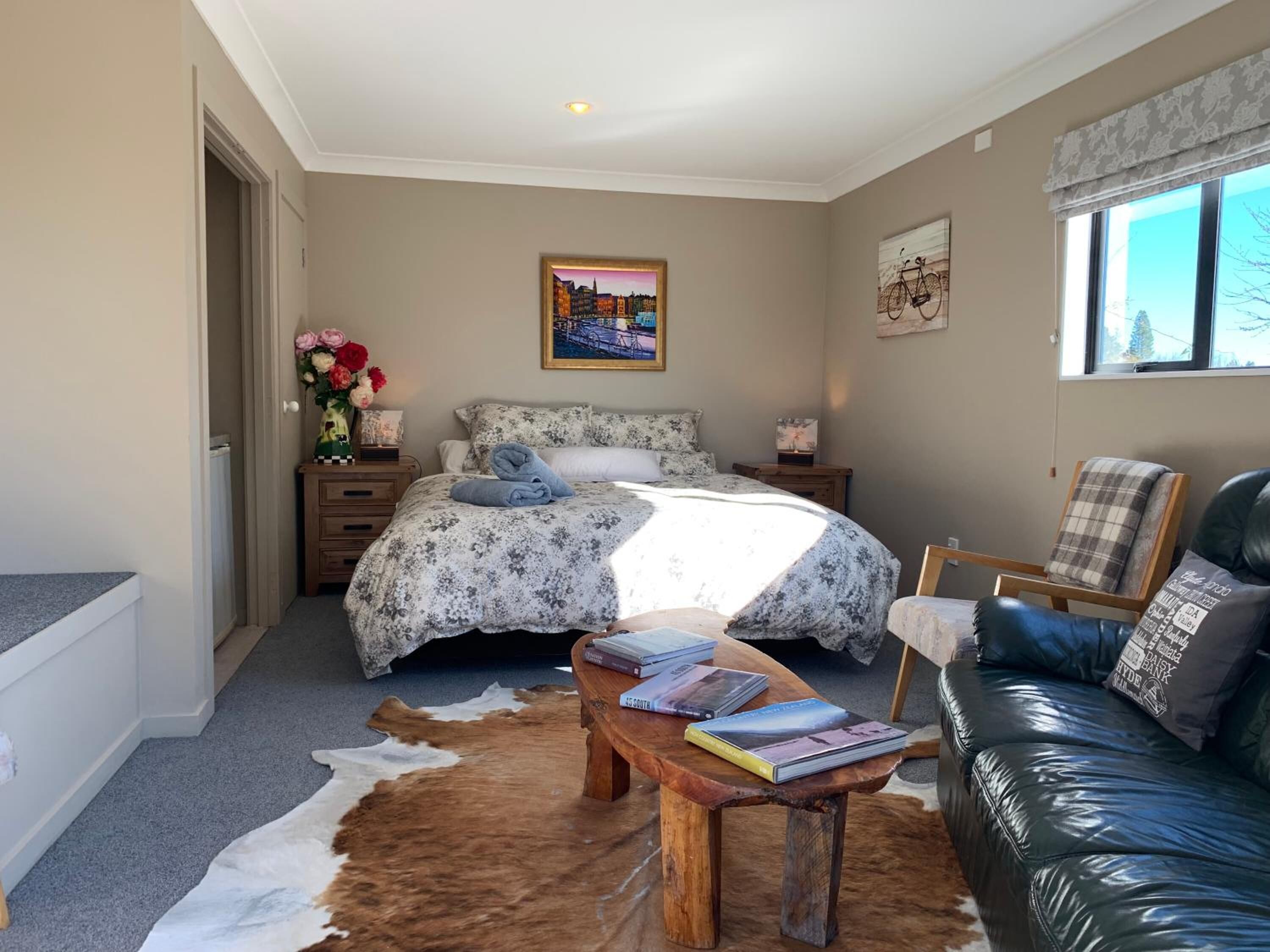 Een slaapkamer met een bed, een houten bijzettafeltje met boeken, een leren bank, een koehuidtapijt en een stadslandschap aan de muur.