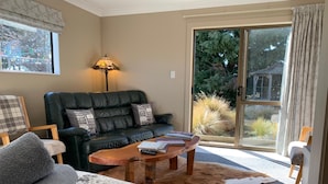 A cozy living room with a leather sofa, a wooden coffee table, and a large window with a view of a garden. - Trout n Trail BnB (Ranfurly)