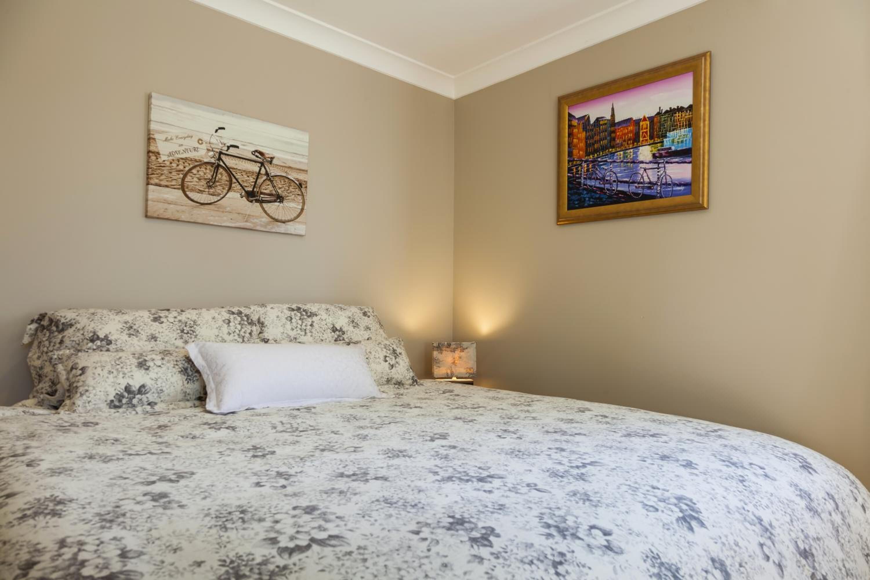 A bedroom with a bed, floral bedding, a white pillow, a wall-mounted bicycle artwork, and a framed cityscape painting.