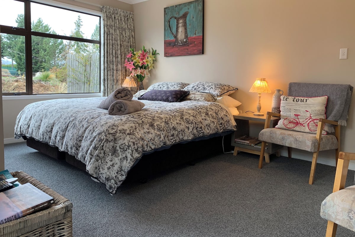 A bedroom with a large bed, a chair, a nightstand, a lamp, a painting, and a window with curtains.