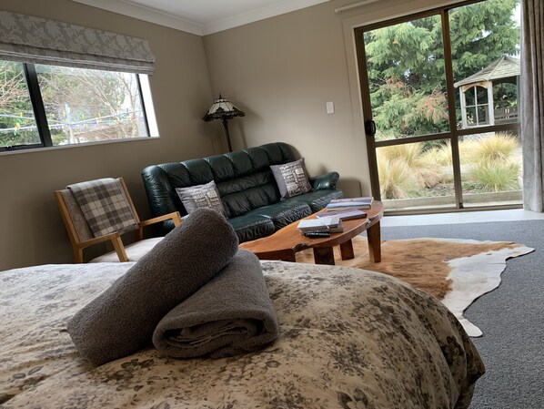 A cozy room with a bed, a leather sofa, a wooden coffee table, and a large window offering a view of greenery outside.