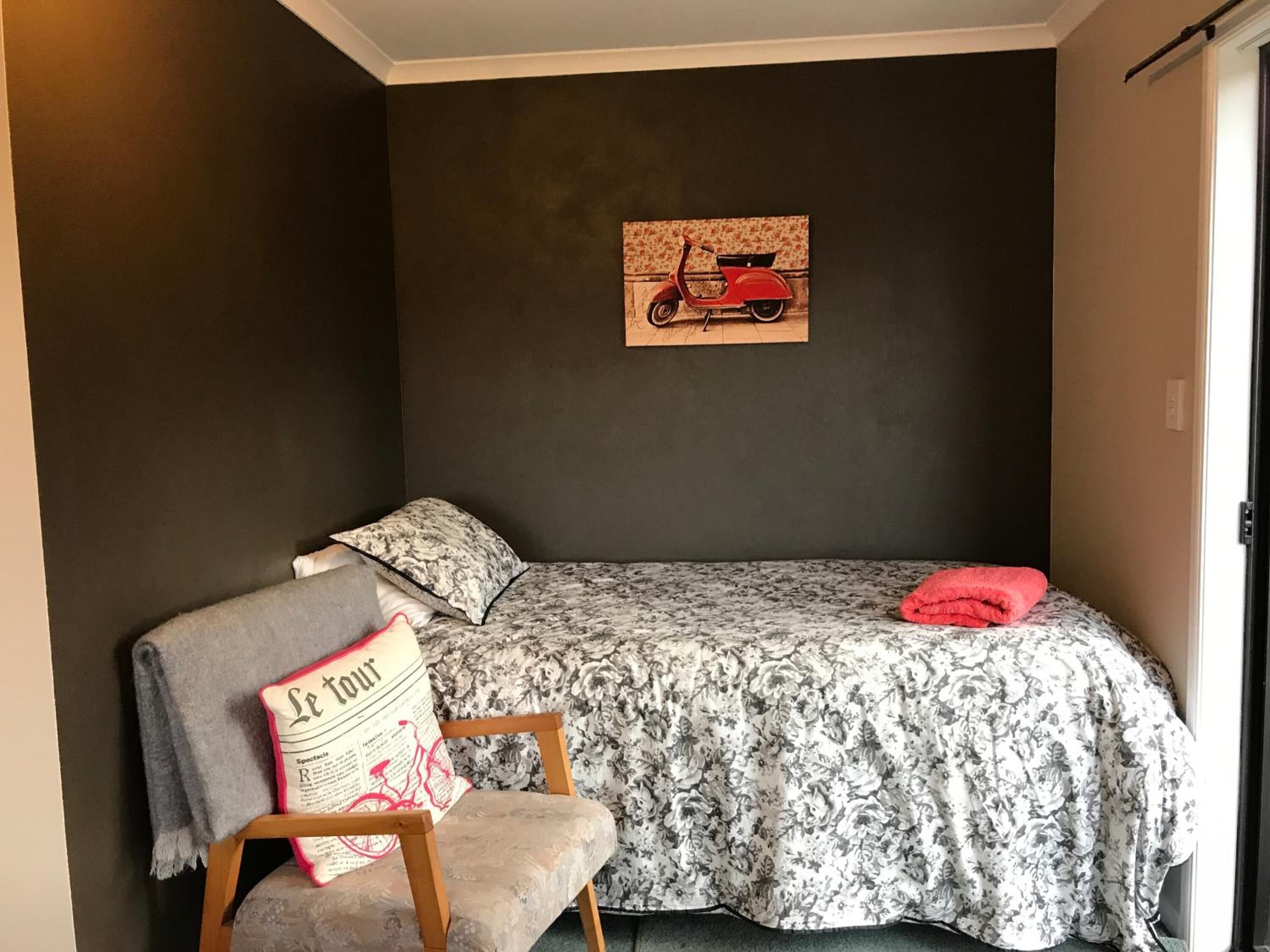 A small, cozy room with a single bed, a chair, and a wall-mounted painting of a scooter.