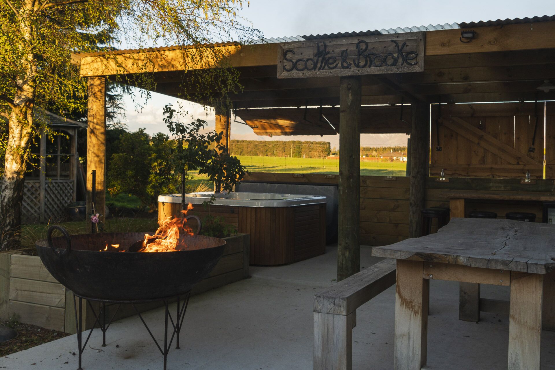 Un espacio al aire libre con jacuzzi, una fogata y una pérgola de madera con un cartel que dice "Scatche & Broke".