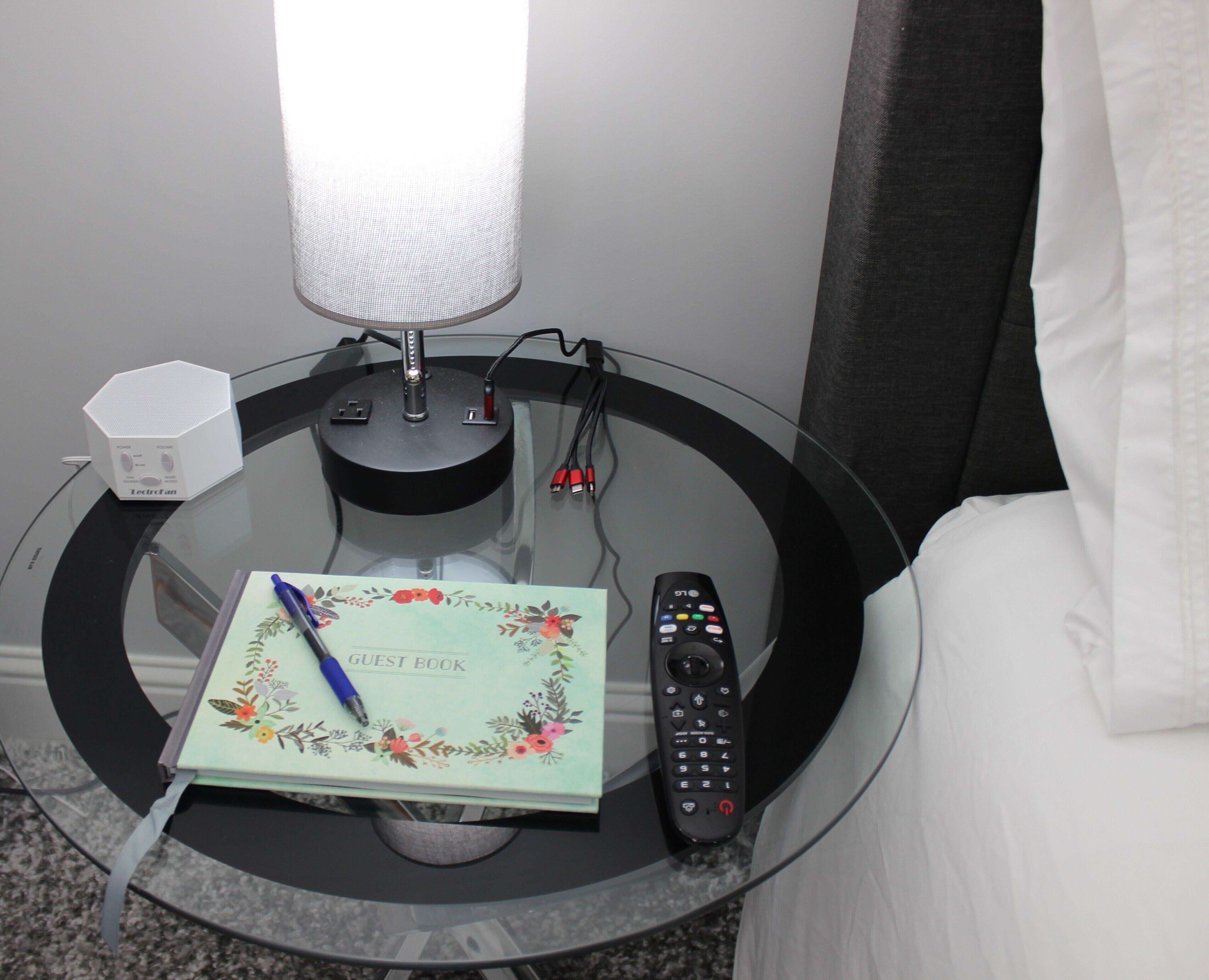 Una mesa de vidrio con un control remoto, un bolígrafo, un cuaderno y una lámpara.