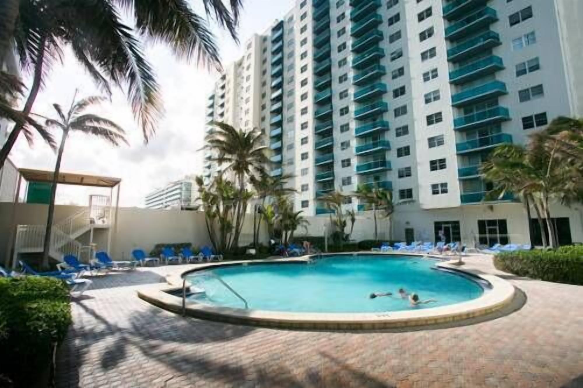 A swimming pool surrounded by lounge chairs and palm trees, with a high-rise building in the background.