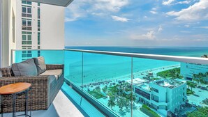 Ein Balkon mit einer Korbuschlaube und einem kleinen Tisch, mit Blick auf einen Strand mit klarem türkisblauem Wasser und ein Resort im Hintergrund.
