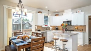 A modern kitchen with white cabinetry, a central island with stools, and a dining area with a wooden table and chairs. - Bluebird Day - Wild River Getaway, Escape to this cozy 2-bedroom retreat in Bend, OR. (Bend)