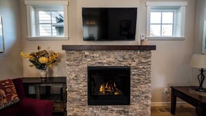 A living room with a stone fireplace, a flat-screen TV mounted above it, and two windows on either side. - Bluebird Day - Wild River Getaway, Escape to this cozy 2-bedroom retreat in Bend, OR. (Bend)