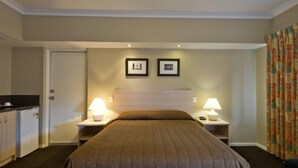 A hotel room with a bed, two bedside lamps, a door, a small table, and a wall with two framed pictures. - Motel Le Grande (Orana)