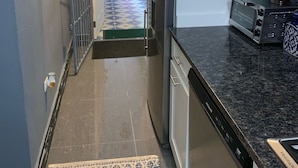 Eine Küche mit Kühlschrank, Geschirrspüler und Toaster. Auf dem Boden liegt ein Teppich mit Muster, und ein Metallgitter führt in einen Außenbereich.