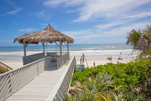 Een houten looppad dat naar een strand leidt, met een rieten hut, palmbomen en parasols in het zand.