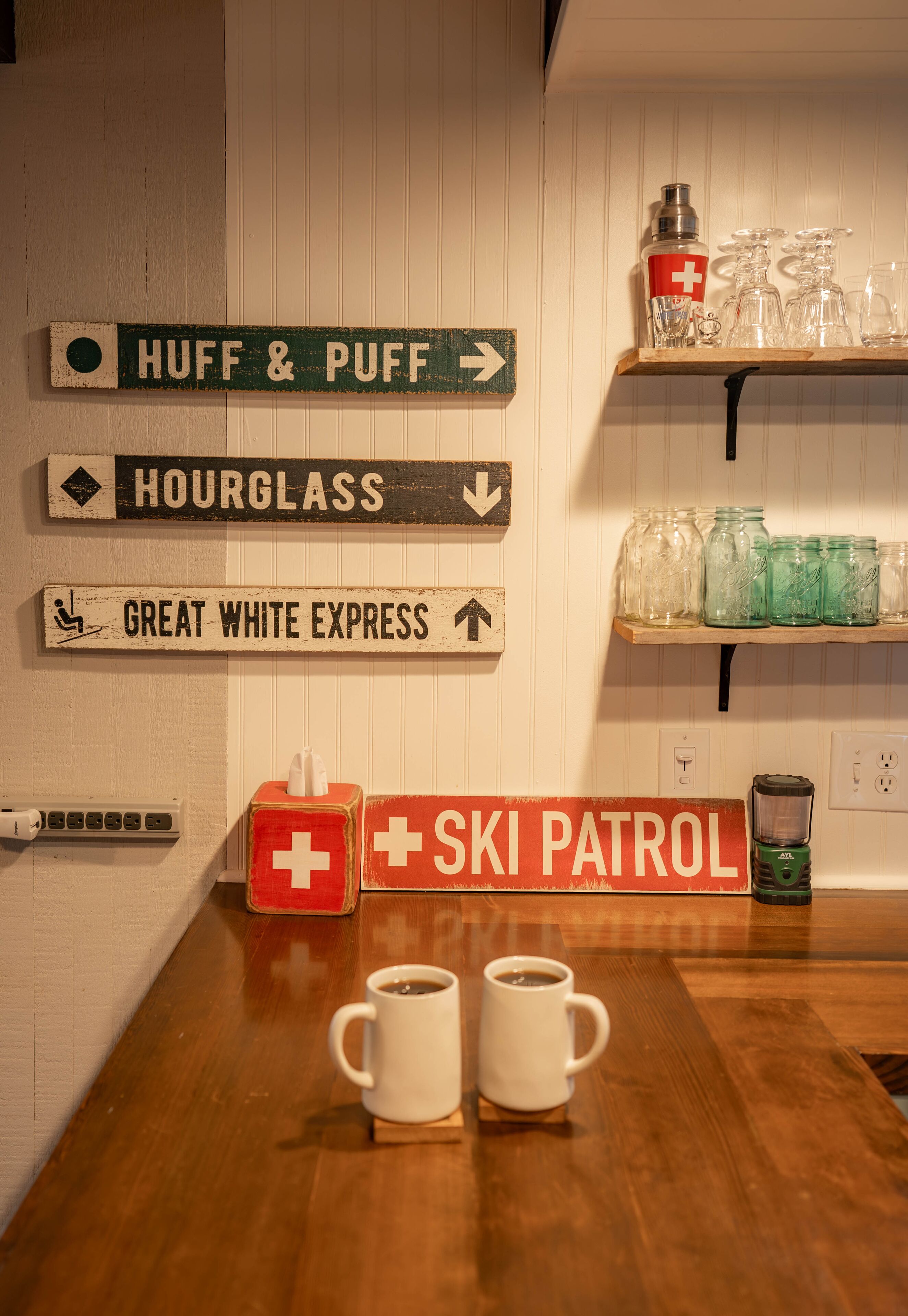 A kitchen area with wooden signs indicating directions to different coffee shops, a red Swiss flag sign, and two white mugs on the counter.