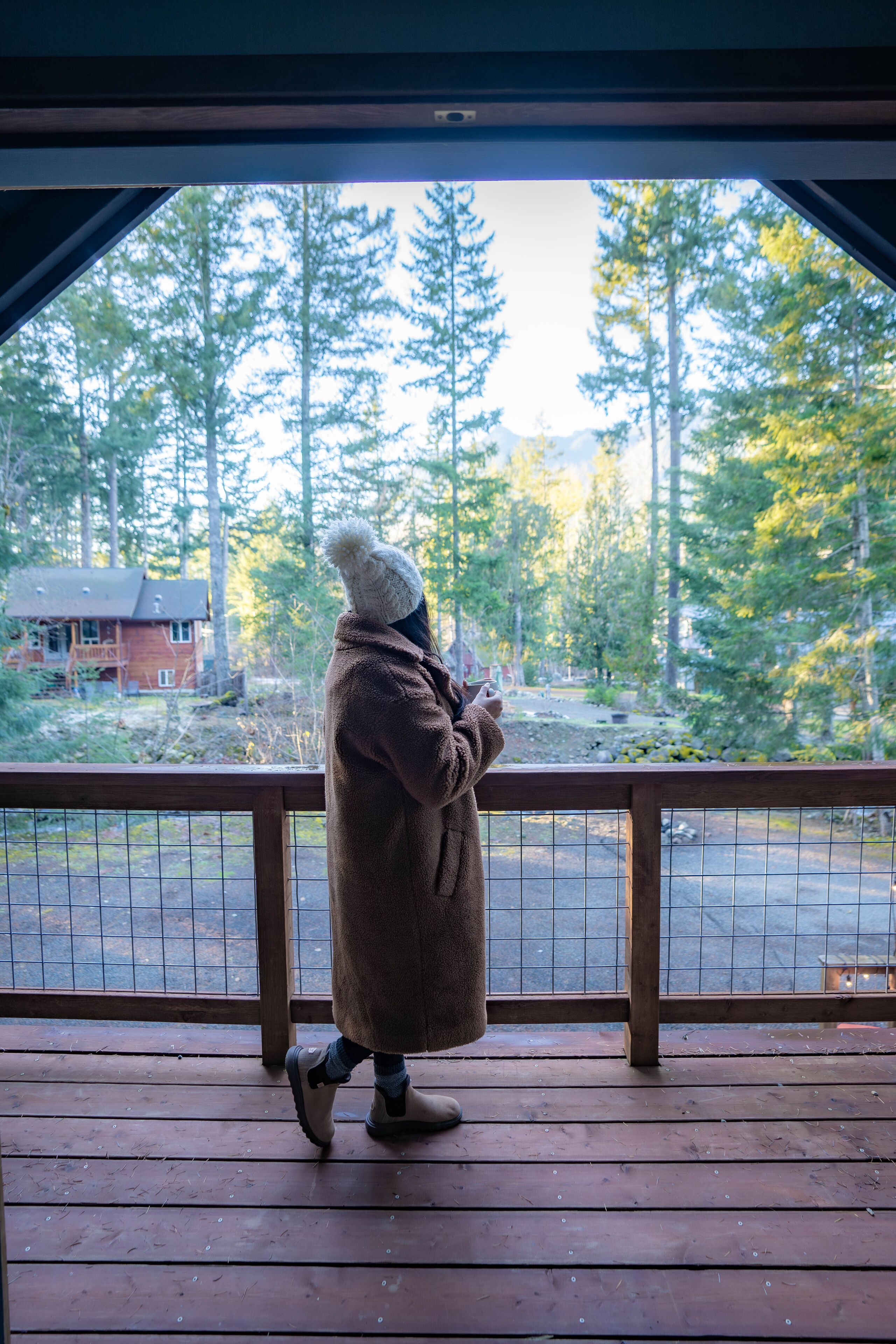 A person standing on a wooden deck, looking out at a forested area with a cabin in the background.