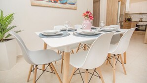 Un comedor con una mesa blanca para cuatro personas, rodeada de sillas blancas, una planta en maceta y un cuadro de barcos en la pared.