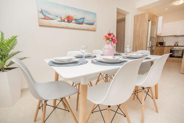 A dining area with a white table set for four, surrounded by white chairs, a potted plant, and a painting of boats on the wall. - Rhodes Marine New House is located in the city of Rhodes! (Rodos)
